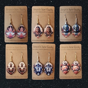 May include: Six pairs of dangle earrings featuring pop culture characters from Star Wars. The earrings are made of wood and have a floral design. The characters include Princess Leia, Chewbacca, Boba Fett, C-3PO, R2-D2, and BB-8.