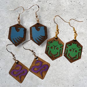 May include: Three pairs of wooden earrings with different designs. The first pair features a blue bird, the second pair features a green bug, and the third pair features a purple snake.