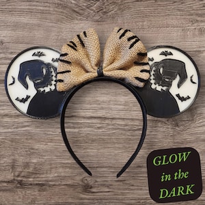 K&ouml;nnte beinhalten: Schwarz-wei&szlig;e, im Dunkeln leuchtende Mickey-Maus-Ohren mit einer Schleife aus Sackleinen. Die Ohren zeigen eine Silhouette von Jack Skellington aus Nightmare Before Christmas.