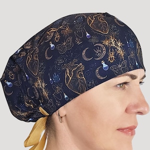 May include: A dark blue scrub cap featuring a repeating pattern of anatomical hearts, crescent moons, moths, beetles, and small potion bottles in gold and blue tones, finished with a solid mustard yellow ribbon tie at the back for adjustment.