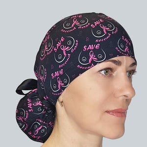 May include: A black scrub cap featuring a repeating pattern of white line-art breast outlines, pink breast cancer awareness ribbons, and the text "SAVE second base" in pink lettering. The cap includes a black ribbon tie at the back.