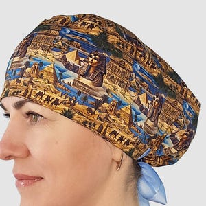 May include: A patterned scrub cap featuring an Egyptian theme. The cap displays pyramids, the Sphinx, camels, and other Egyptian imagery in shades of brown, blue, and gold. A light blue ribbon is tied at the back.