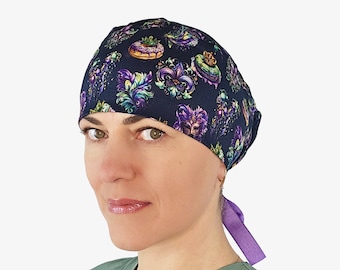 May include: A navy blue surgical scrub cap featuring a colorful Mardi Gras design. The pattern includes fleur-de-lis, masks, and decorative elements in purple, green, and gold. The cap has a purple tie in the back.