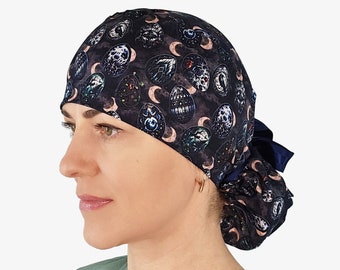 May include: A dark blue surgical scrub cap with a repeating pattern of stylized skulls, moons, and decorative elements. The cap is tied in a ponytail style with a dark blue ribbon. The background is plain white.
