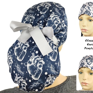 May include: A navy blue surgical scrub cap with a white anatomical heart and celestial pattern. The cap has a gray ribbon bow and is designed for a ponytail style. The text "Classic Euro Ponytail" is also visible.