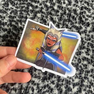 May include: A sticker featuring a cartoon illustration of a female character from Star Wars. The character is wielding two blue lightsabers and has a fierce expression on her face. The background is a fiery orange and yellow.