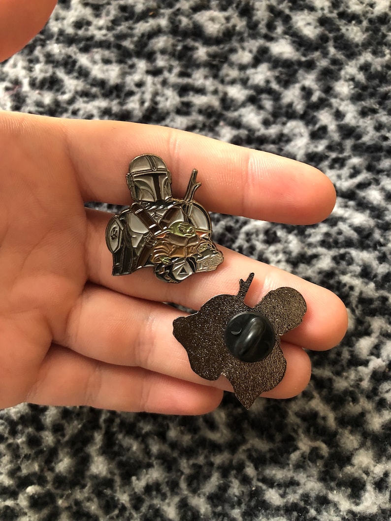 K&ouml;nnte beinhalten: Eine goldfarbene Metallsticker mit einer Figur aus dem Star Wars-Franchise, The Mandalorian, der ein kleines gr&uuml;nes Alien, bekannt als Grogu, in seinen Armen h&auml;lt. Die Anstecknadel hat ein detailliertes Design und eine schwarze, schmetterlingsf&ouml;rmige R&uuml;ckseite.