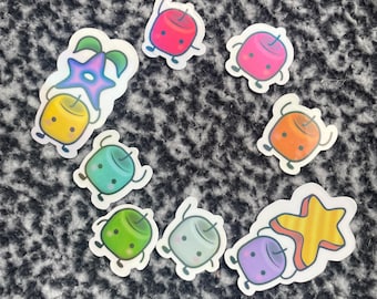 May include: A selection of colourful cartoon stickers. The stickers depict marshmallow-like characters in red, pink, orange, green, and purple. Some have star and leaf accents. The stickers are arranged on a textured, dark grey surface.