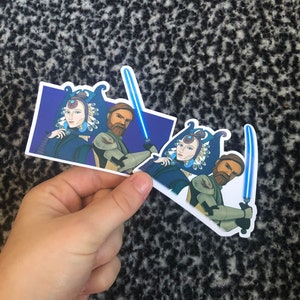 May include: Two sticker sets featuring a blue and white illustration of a woman with a crown and a man with a beard. Both are holding blue lightsabers.