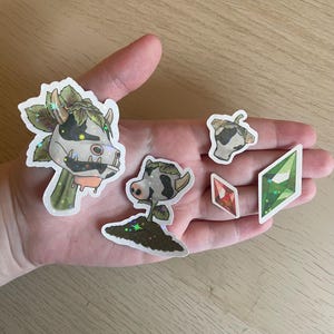 以下が含まれることがあります： 牛をテーマにしたデザインのホログラフィックステッカーのコレクション。ステッカーには、葉っぱのついた牛の頭、牛の頭、土から生える牛、赤と緑の宝石型のステッカー2枚が含まれています。