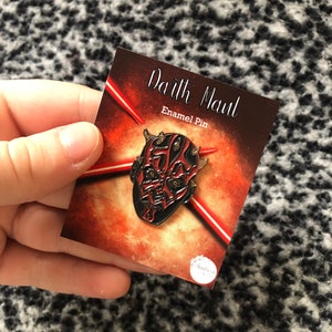 Peut inclure: Une épingle en émail rouge et noire représentant le visage de Dark Maul. L'épingle est sur une carte avec le texte "Darth Maul Enamel Pin".