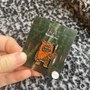 Könnte beinhalten: Ein Ewok-Emaille-Pin auf grünem Hintergrund. Der Pin zeigt einen braunen Ewok-Charakter, der einen Speer hält, mit einer orangefarbenen Kapuze. Der Pin befindet sich auf einer Karte mit der Aufschrift "Ewok Enamel Pin".