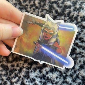 May include: A sticker featuring a character from Star Wars, Ahsoka Tano, wielding two blue lightsabers. The character has orange skin, white and blue markings, and a determined expression. The background is a blend of orange and yellow.