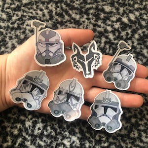 May include: A set of seven stickers featuring Star Wars clone troopers. The stickers are in a grey and white colour scheme and feature different designs, including a wolf head and a clone trooper helmet.