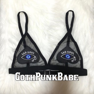 Puede incluir: Sujetador triangular de malla negra con dos parches negros en forma de corazón. Cada parche tiene un ojo turco azul y el texto "SAD SOUL" en blanco.