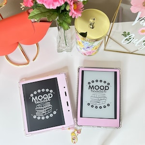 Könnte beinhalten: Zwei rosa E-Reader-Hüllen mit klaren Abdeckungen. Die Hüllen haben eine schwarz-weiße Grafik eines Stapels Bücher mit dem Text "MOOD reader" über den Büchern. Die Hüllen befinden sich auf einer weißen Oberfläche.