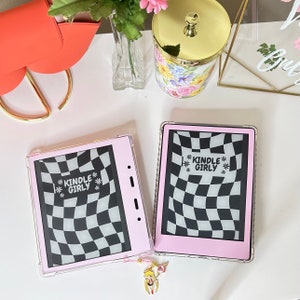 Puede incluir: Dos lectores electrónicos Kindle rosas con fundas transparentes, cada uno con un patrón de cuadros en blanco y negro y el texto "KINDLE GIRLY". Uno tiene un dije. El fondo incluye un bolso coral, flores y un tarro decorativo.