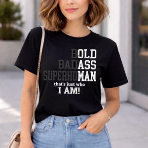 Puede incluir: Camiseta negra con las palabras "BOLD BADASS SUPERHUMAN" en un diseño en capas. Debajo de las palabras está la frase "that's just who I AM!" en blanco. La camiseta es de cuello redondo.