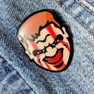 May include: An enamel pin featuring a cartoon face with a sinister smile. The face has red stripes and a black outline.
