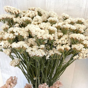 May include: A bouquet of dried white statice flowers with green stems. The flowers are arranged in a vase and are in focus. The background is a white wooden surface.