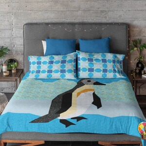 Puede incluir: Un edredón estampado en azul y blanco con un diseño de pingüino. El edredón está en una cama con cabecera gris y dos almohadas azules.