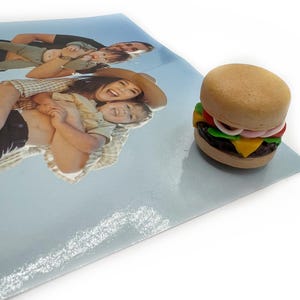 Peut inclure: Un mini-burger avec un pain aux graines de s&eacute;same, de la laitue, de la tomate, de l'oignon, du fromage et une galette fonc&eacute;e. Le burger est pos&eacute; sur une surface bleu clair &agrave; c&ocirc;t&eacute; d'une photo de famille.