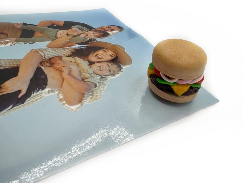 Peut inclure: Un mini-burger avec un pain aux graines de s&eacute;same, de la laitue, de la tomate, de l'oignon, du fromage et une galette fonc&eacute;e. Le burger est pos&eacute; sur une surface bleu clair &agrave; c&ocirc;t&eacute; d'une photo de famille.