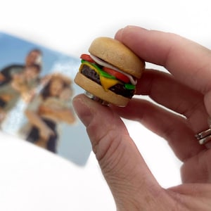Peut inclure: Bague miniature en forme de hamburger avec pain, steak, fromage, tomate, laitue et oignon. La bague est tenue devant un arri&egrave;re-plan flou. Un accessoire amusant.