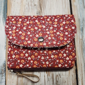 Peut inclure: Une petite pochette en tissu matelassé avec un fond rouge foncé orné d'un motif répétitif de minuscules fleurs jaunes et blanches et de feuilles vertes. La pochette possède un rabat arrondi fixé par un bouton-pression argenté et une fermeture éclair.