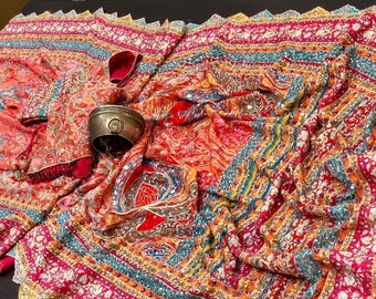 May include: An ornate textile display featuring a vibrant array of colors including red, orange, and teal. The fabric is intricately patterned with detailed designs and embellished with gold trim. A small metal container and a red accessory are also present.