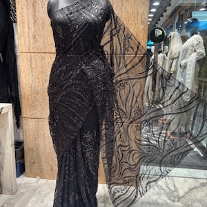 May include: A black one-shoulder gown featuring intricate sequin embroidery in a swirling leaf pattern. The garment includes a sheer, flowing panel extending from the shoulder, showcasing a detailed black threadwork design across the fabric.