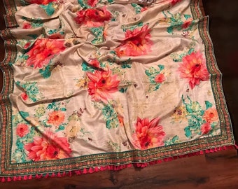 May include: A cream-colored silk saree with a floral print in shades of pink, green, and yellow. The saree has a green and gold border with pink tassels.