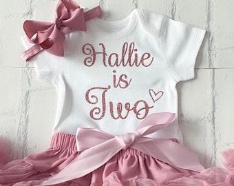 May include: A white onesie with the text "Hallie is Two" in rose gold glitter. The onesie is paired with a pink tulle tutu and a pink bow headband.