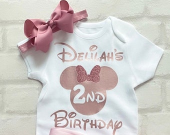 May include: A white onesie with a rose gold Minnie Mouse design, the text "Delilah's 2nd Birthday", and a rose gold bow headband. A pink tulle skirt with a rose gold bow is also included.