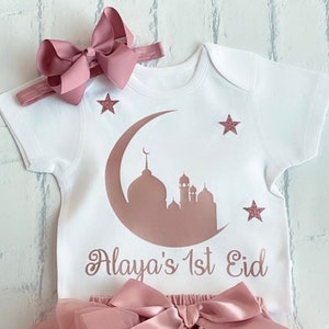 May include: A white baby outfit with a pink bow headband and a matching tutu. The onesie features a mosque and crescent moon design with the text "Alaya's 1st Eid". Pink glitter stars are also on the onesie.