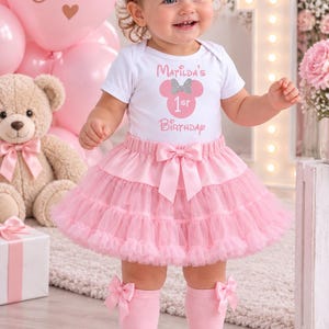 May include: A white short-sleeved bodysuit with the text Matilda's 1st Birthday and a pink mouse silhouette, paired with a layered pink tulle tutu skirt featuring a satin bow, a matching pink headband with a bow, and pink knee-high socks with bows.