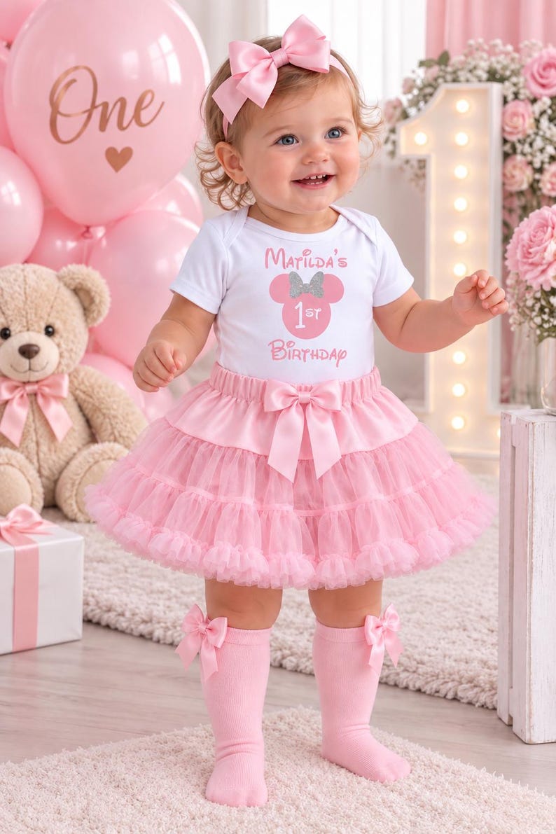 May include: A white short-sleeved bodysuit with the text Matilda's 1st Birthday and a pink mouse silhouette, paired with a layered pink tulle tutu skirt featuring a satin bow, a matching pink headband with a bow, and pink knee-high socks with bows.