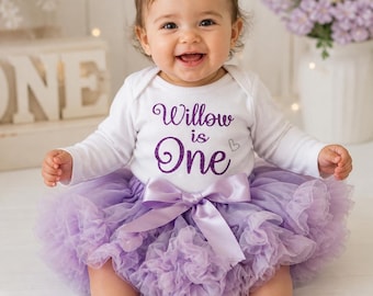 May include: A baby wearing a white long-sleeved onesie with the text "Willow is One" in purple glitter, paired with a lavender tulle skirt and a matching satin bow. The baby is smiling.