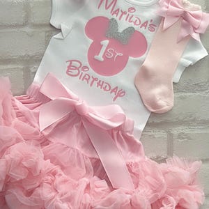 May include: A white short-sleeved bodysuit with pink text reading Matilda's 1st Birthday and a pink mouse silhouette with a silver glitter bow. Includes a pink ruffled tutu skirt with a large satin bow, a pink headband, and pink socks with bows.