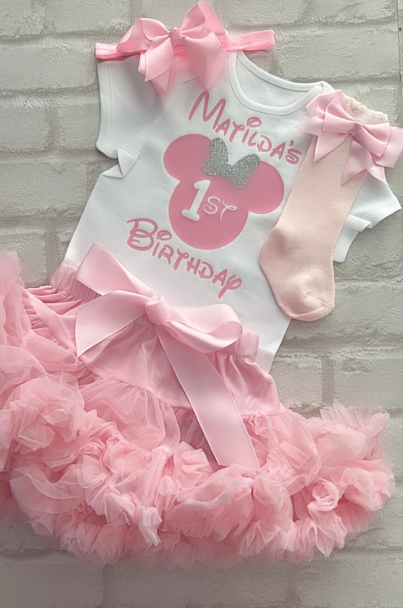 May include: A white short-sleeved bodysuit with pink text reading Matilda's 1st Birthday and a pink mouse silhouette with a silver glitter bow. Includes a pink ruffled tutu skirt with a large satin bow, a pink headband, and pink socks with bows.