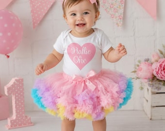 May include: A white short-sleeved bodysuit featuring a pink heart graphic with the text Hallie is One, paired with a multi-colored pastel tulle tutu skirt with a pink satin bow at the waist and a matching pink satin bow headband.