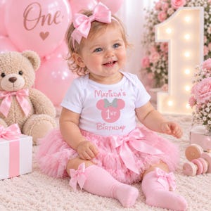 May include: A white short-sleeved bodysuit with the text Matilda's 1st Birthday and a pink mouse ear graphic, paired with a pink ruffled tulle tutu skirt, a matching pink satin hair bow headband, and pink knee-high socks with small bows.