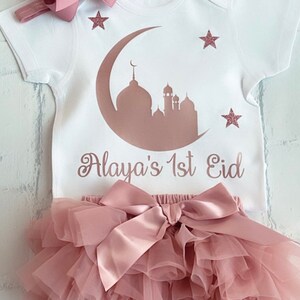 May include: A white baby outfit set with a matching headband. The onesie features a mosque silhouette, crescent moon, and stars in a rose gold color, with the text "Alaya's 1st Eid". The set includes a tutu skirt with a large bow.