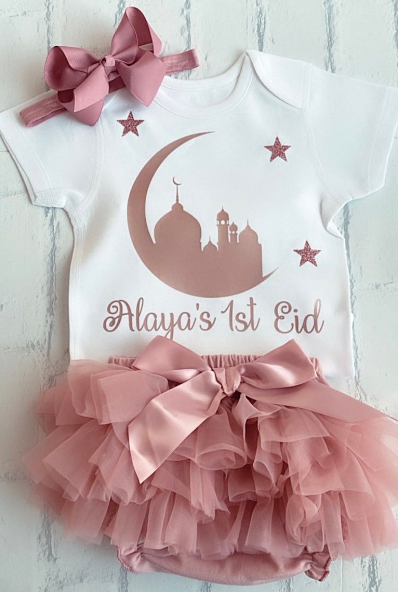 May include: A white baby outfit set with a matching headband. The onesie features a mosque silhouette, crescent moon, and stars in a rose gold color, with the text "Alaya's 1st Eid". The set includes a tutu skirt with a large bow.