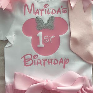 May include: A white short-sleeved bodysuit featuring the pink text Matilda's 1st Birthday and a pink mouse silhouette with a silver glitter bow. The set includes a pair of light pink socks with a pink bow and pink ruffled shorts with a large satin bow.