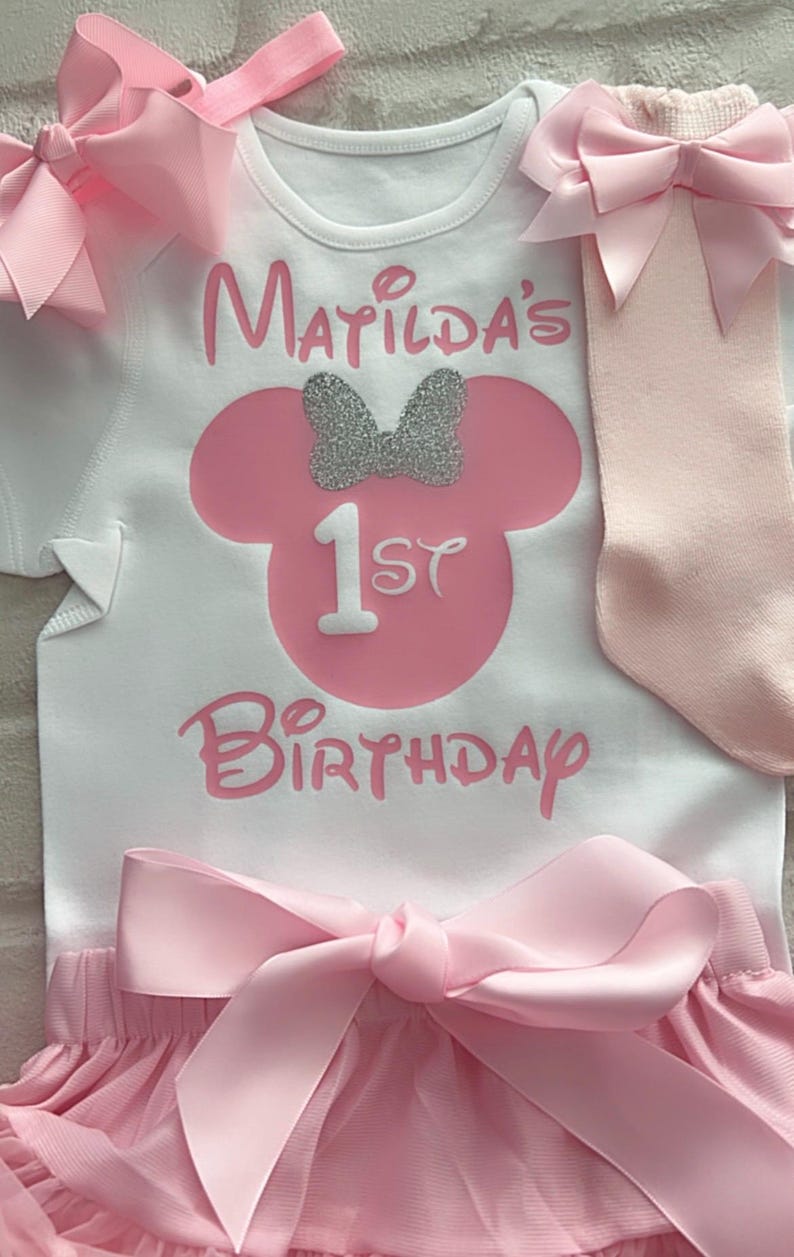 May include: A white short-sleeved bodysuit featuring the pink text Matilda's 1st Birthday and a pink mouse silhouette with a silver glitter bow. The set includes a pair of light pink socks with a pink bow and pink ruffled shorts with a large satin bow.
