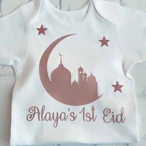 May include: White baby onesie with a rose gold crescent moon and mosque design. The text "Alaya's 1st Eid" is printed below the design, with three rose gold glitter stars.