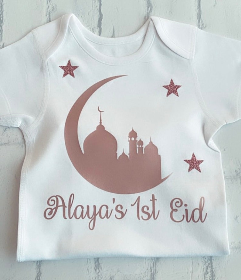 May include: White baby onesie with a rose gold crescent moon and mosque design. The text "Alaya's 1st Eid" is printed below the design, with three rose gold glitter stars.