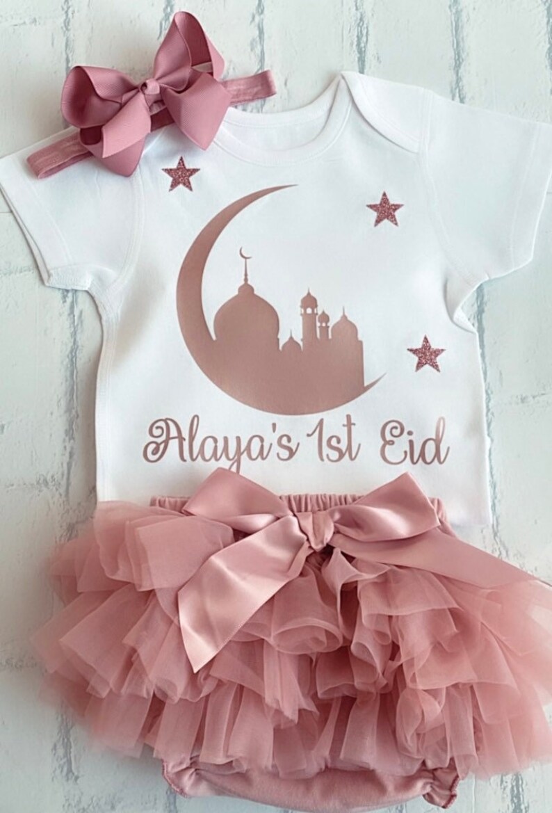 May include: A white baby outfit with a pink bow headband and a matching tutu. The onesie features a mosque and crescent moon design with the text "Alaya's 1st Eid". Pink glitter stars are also on the onesie.