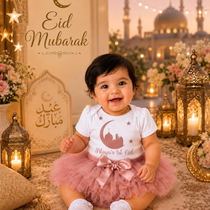 May include: A smiling baby wearing a white onesie with a crescent moon and mosque design, paired with a pink tutu and bow. The text "Maya's 1st Eid" is visible. The background includes lanterns, flowers, and the words "Eid Mubarak".